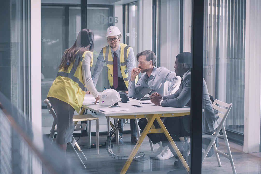 Projektteam mit Warnwesten und Bauhelmen bespricht Pläne und Unterlagen in einem modernen Besprechungsraum.