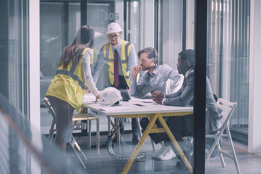 Projektteam mit Warnwesten und Bauhelmen bespricht Pläne und Unterlagen in einem modernen Besprechungsraum.
