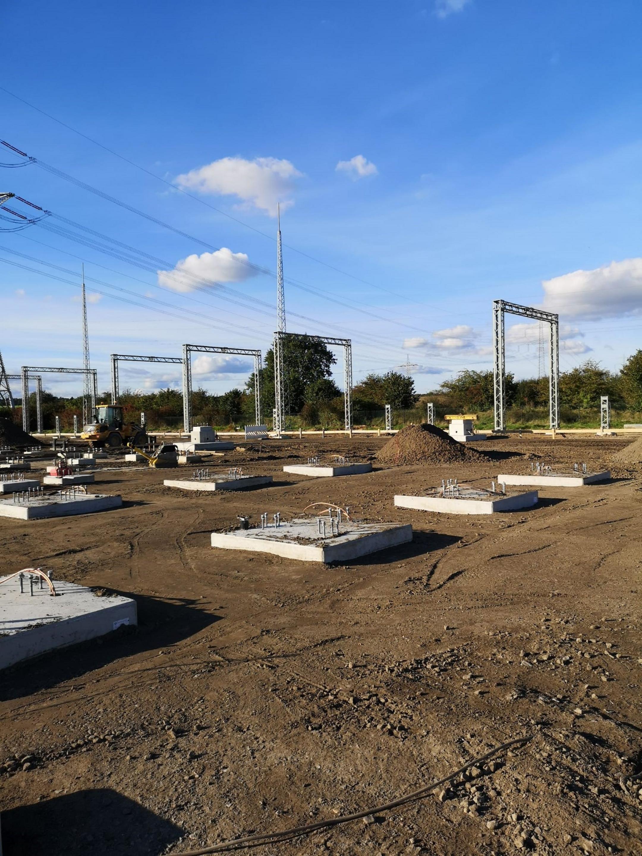 Baustelle mit Betonfundamenten, Metallstrukturen und Strommasten vor klarem Himmel und grüner Umgebung
