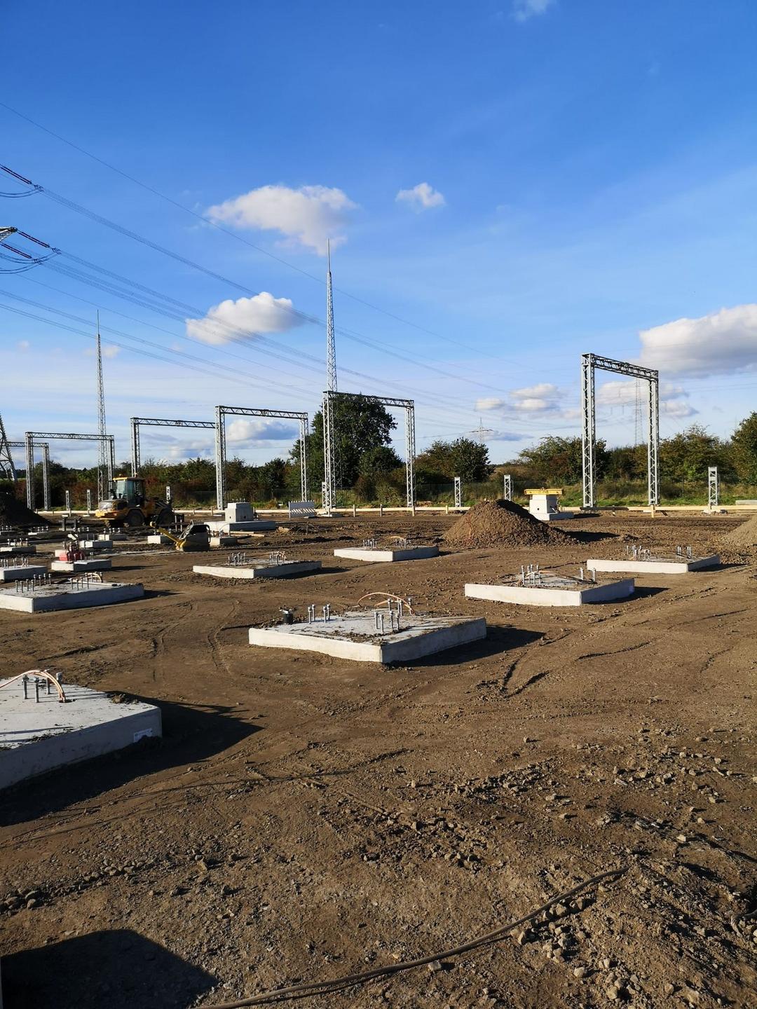 Baustelle mit Betonfundamenten, Metallstrukturen und Strommasten vor klarem Himmel und grüner Umgebung