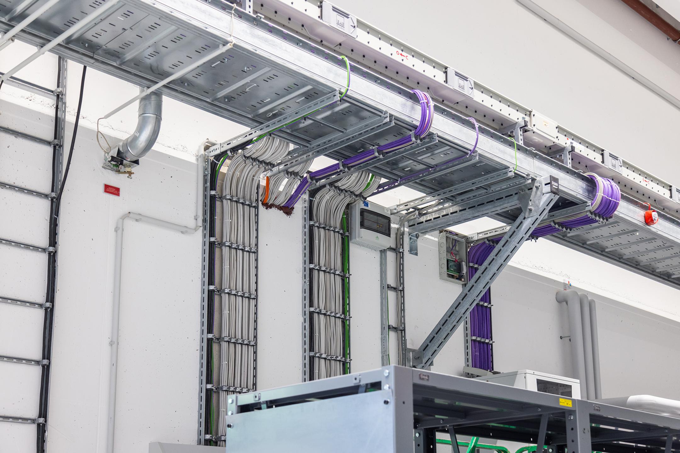 An der Wand angebrachte Kabelleitern mit mehrfarbigen Kabeln und Kabelschellen. Eine Weitspannrinne ist im Anschnitt zu erkennen.