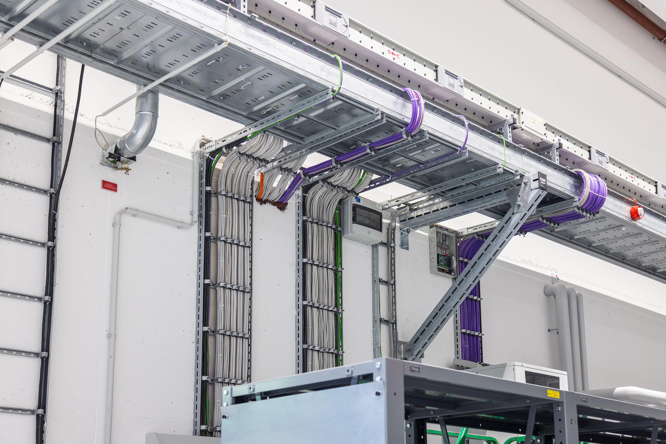 An der Wand angebrachte Kabelleitern mit mehrfarbigen Kabeln und Kabelschellen. Eine Weitspannrinne ist im Anschnitt zu erkennen.