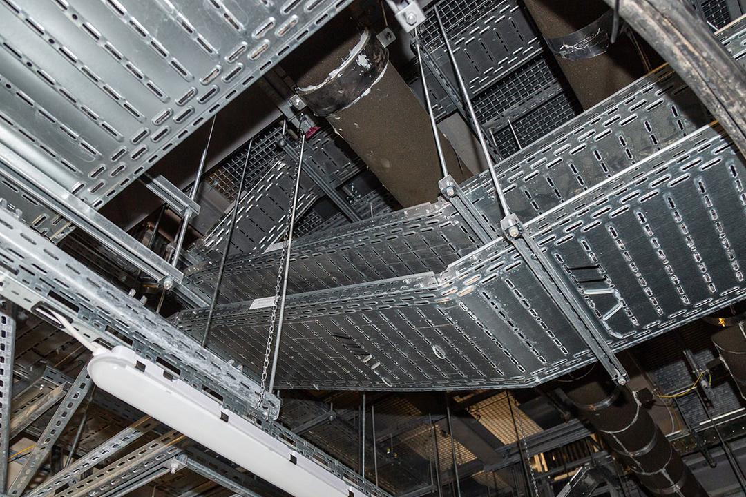 Blick an die Decke auf Kabelrinnen und Rohrleitungen, teils mit Isolierung, in einem Technikraum