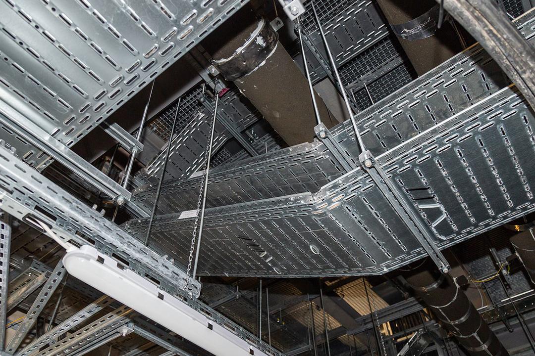 Blick an die Decke auf Kabelrinnen und Rohrleitungen, teils mit Isolierung, in einem Technikraum