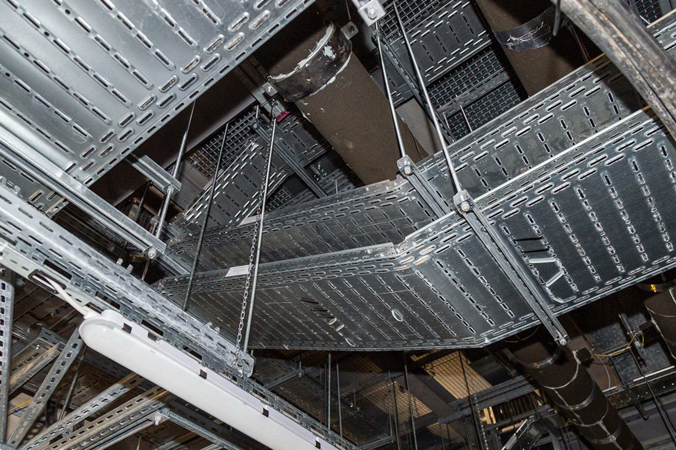 Blick an die Decke auf Kabelrinnen und Rohrleitungen, teils mit Isolierung, in einem Technikraum