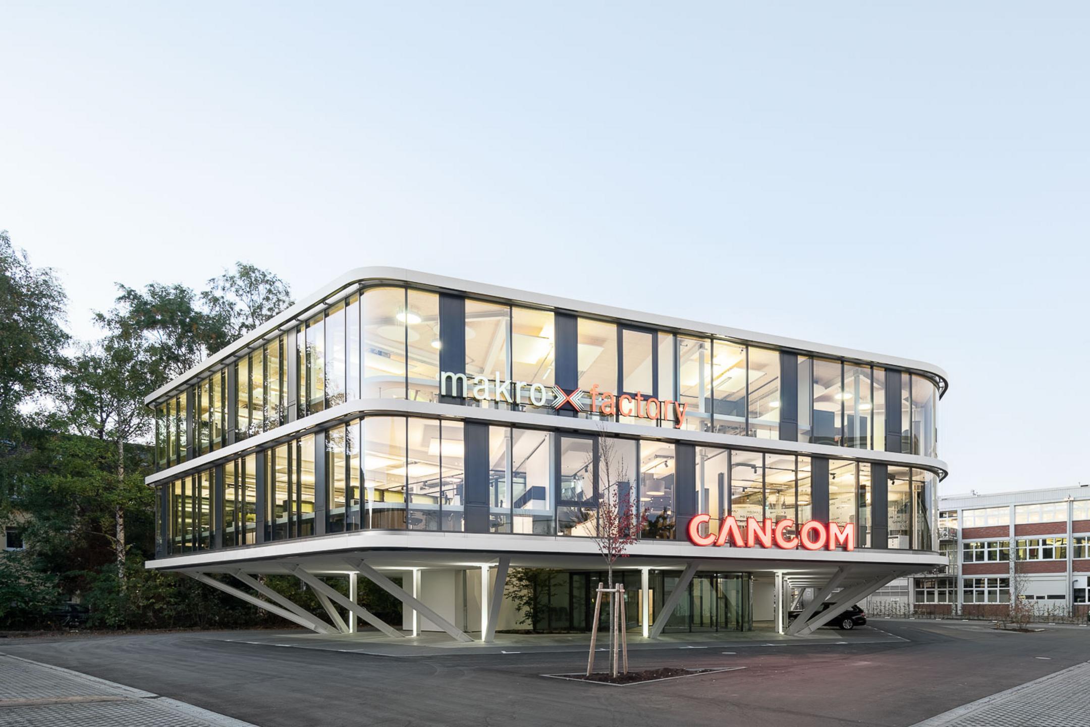 Gebäudeansicht frontal von draußen, Bäume im Hintergrund, blauer Himmel. Dämmerung