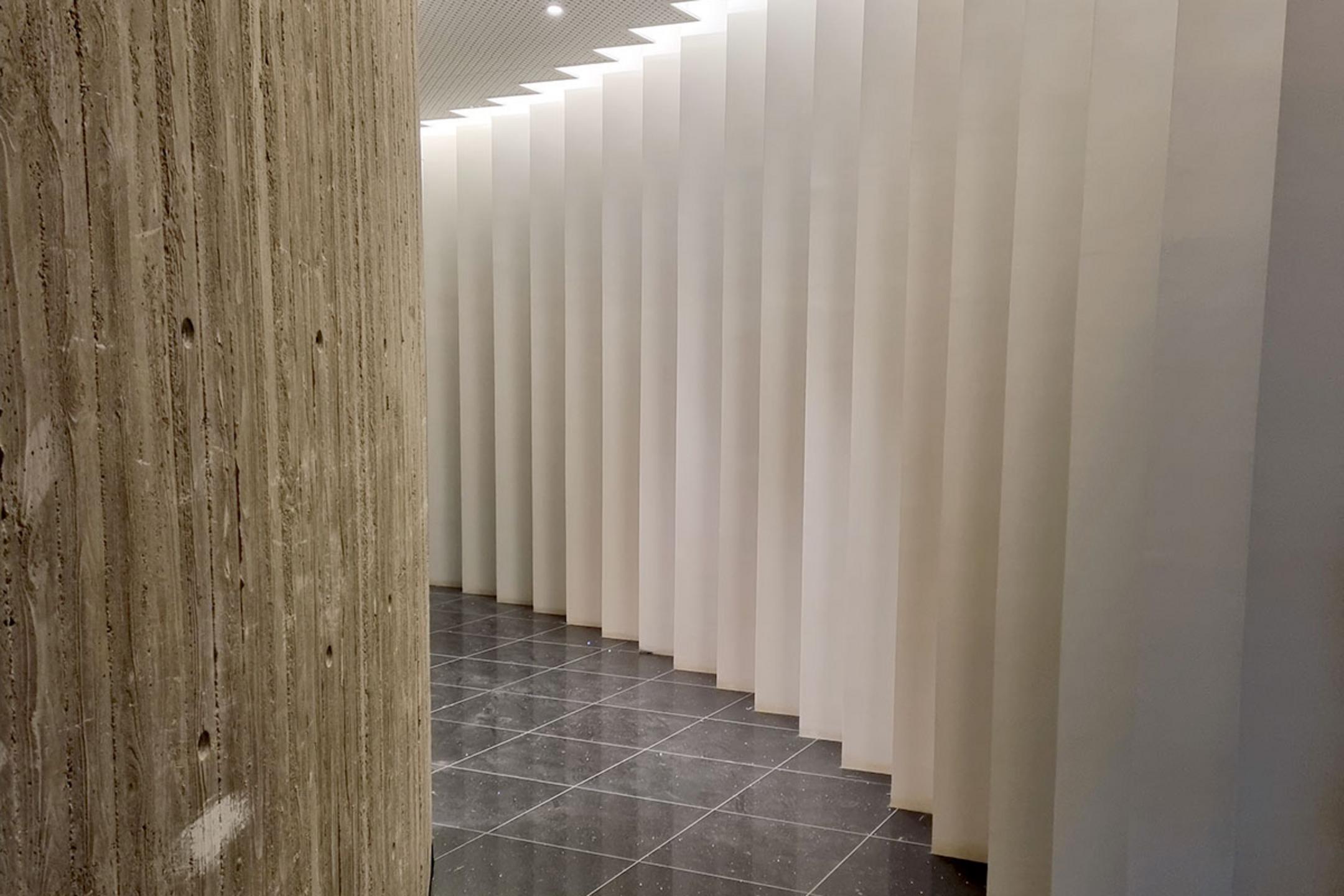 Gebogener Sichtbeton links, rechts weiße vertikale Lamellen, glänzender schwarzer Fliesenboden darunter