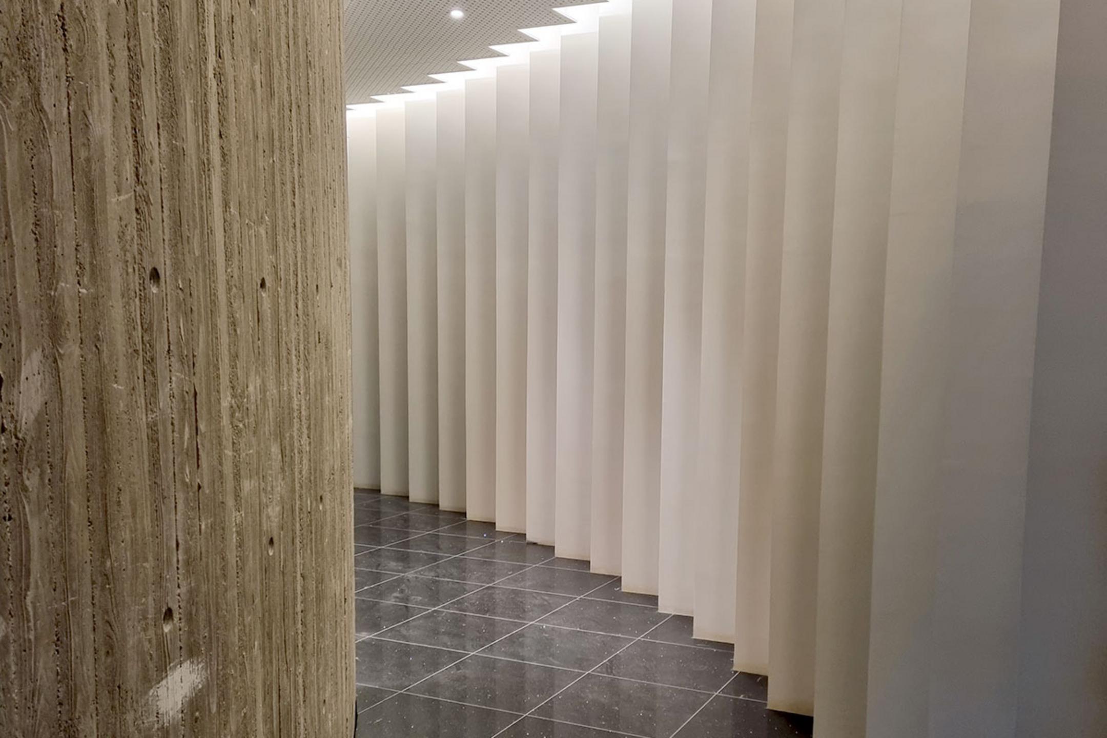Gebogener Sichtbeton links, rechts weiße vertikale Lamellen, glänzender schwarzer Fliesenboden darunter