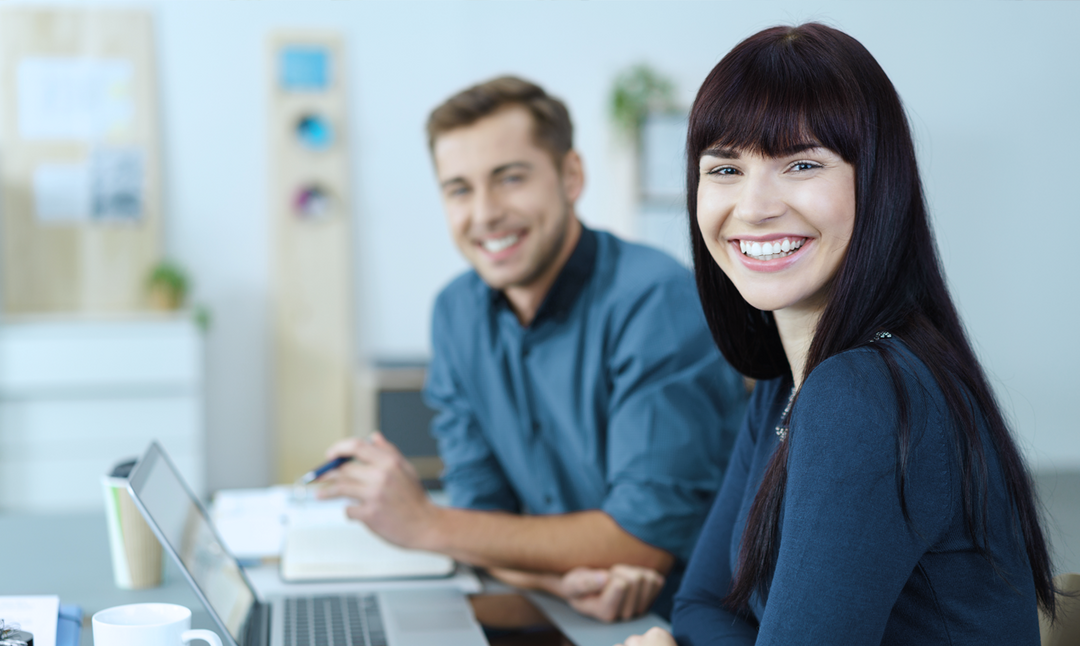 Ein junger Mann und eine junge Frau sitzen in einem Bürosetting lachend an ihren Arbeitsplätzen und blicken in die Kamera.