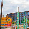 Grün-weiße Schalrohre auf einer Baustelle mit sichtbarer Bewehrung, im Hintergrund ist ein Gebäude mit Fensterfront erkennbar, bewölkter Himmel.