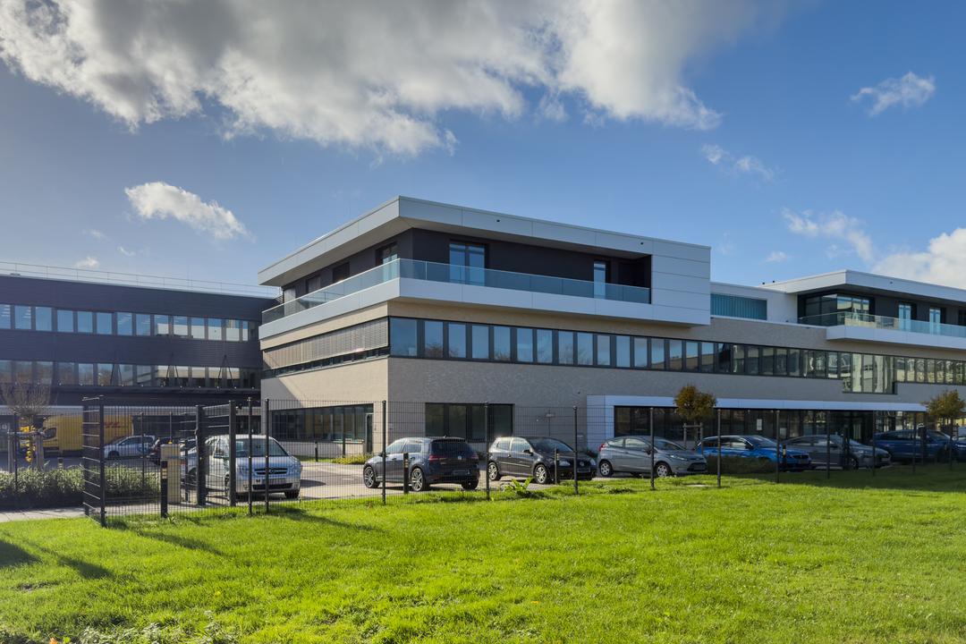 Modernes Bürogebäude mit Glasfassade, Parkplatz und grüner Wiese unter blauem Himmel mit Wolken
