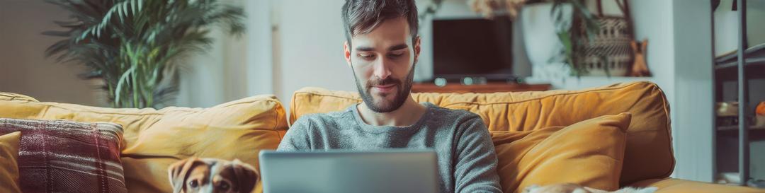 Frontalaufnahme eines jungen Mannes, der auf dem Sofa sitzt und auf seinen Laptop auf seinem Schoß blickt. Links und rechts von ihm liegt ein Hund.