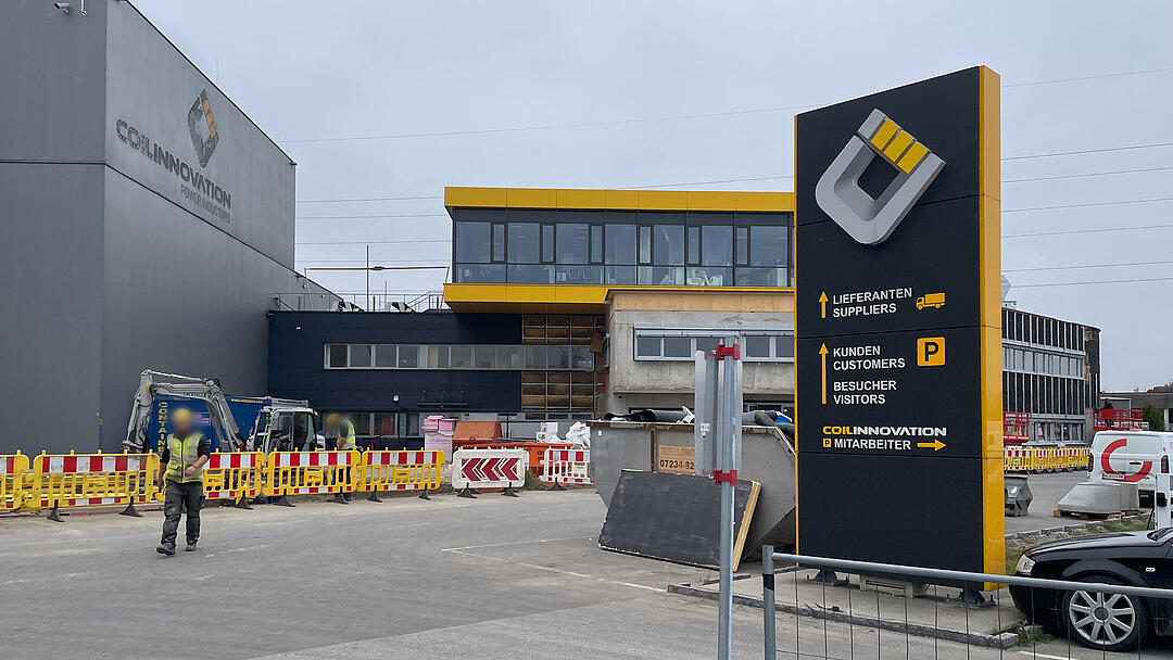 Außenansicht eines Unternehmensstandorts während der Bauphase mit moderner Gebäudefassade, Baustellenabsperrungen, Baufahrzeugen und Wegweiser für Besucher, Kunden und Lieferanten.