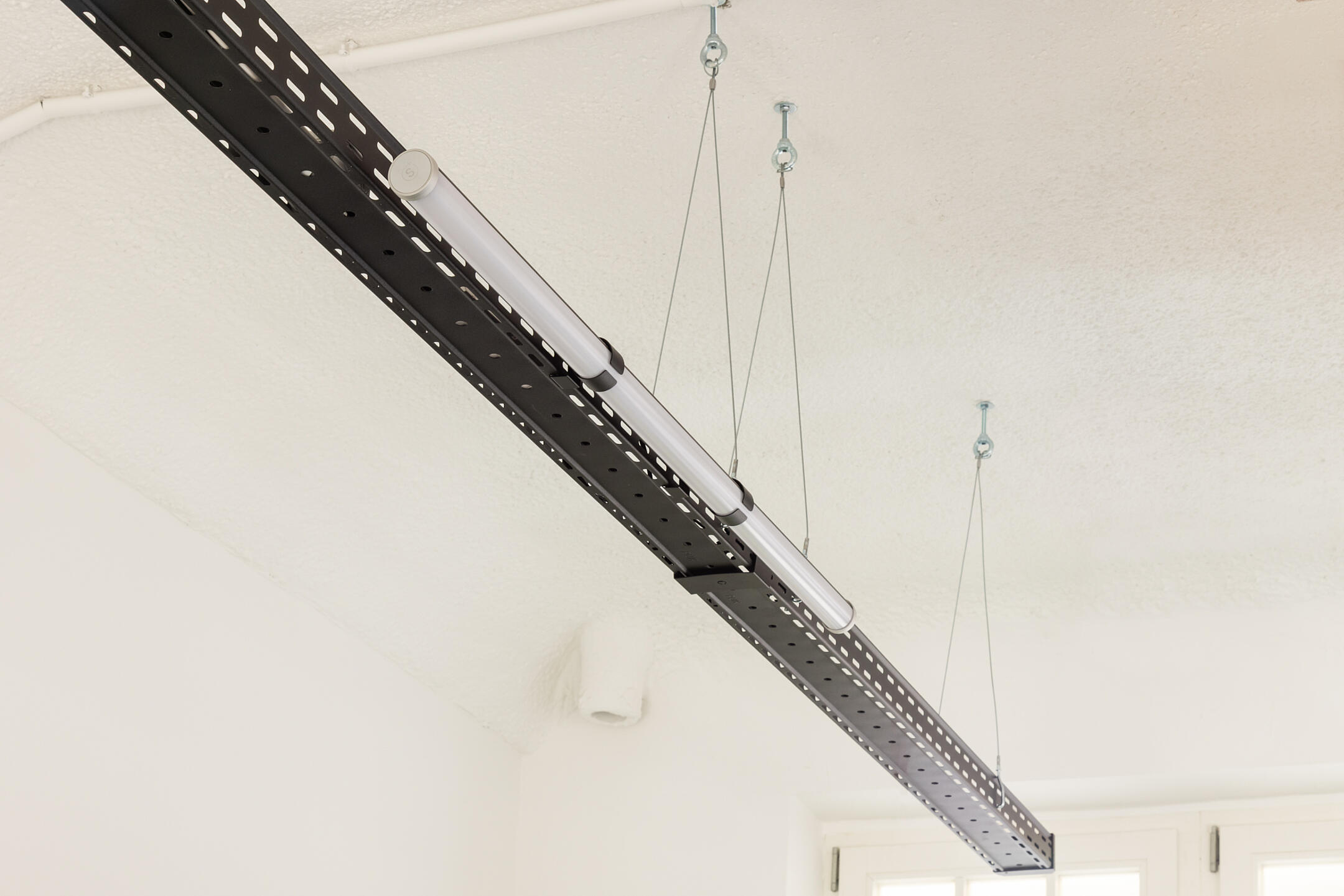 Eine schwarze Kabelrinne hängt von der Decke, daran ist eine schmale runde Leuchte befestigt.