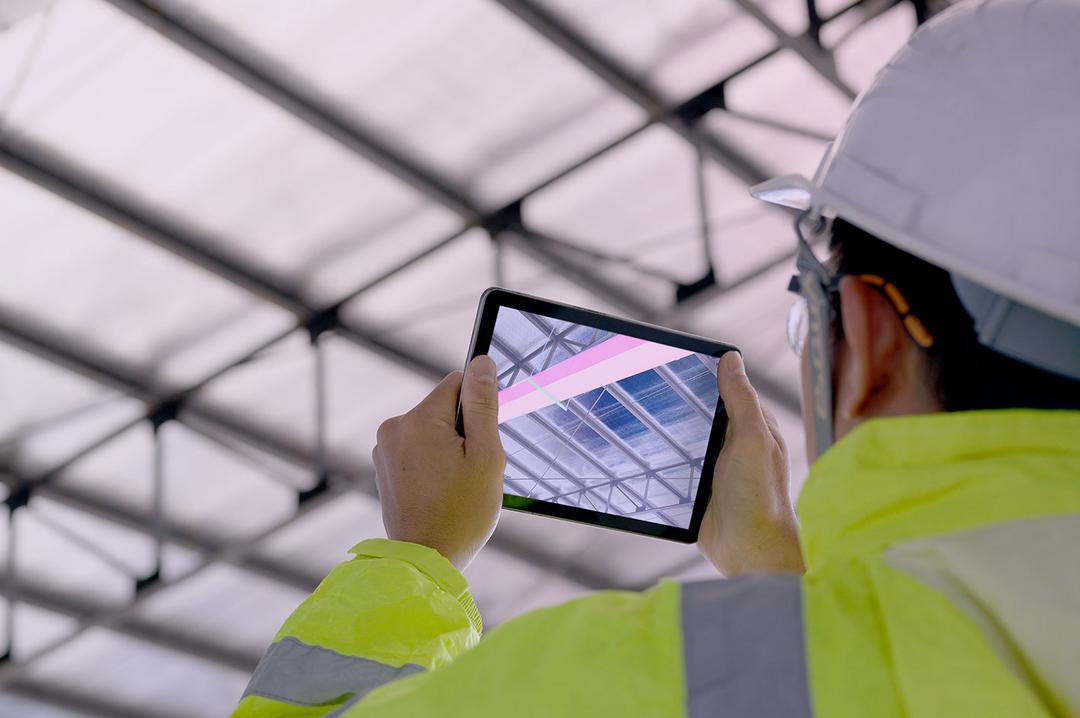 Mann mit Warnweste und Bauhelm blickt auf ein Tablet, auf dem in die Kabelführung in dem Raum simuliert wird.