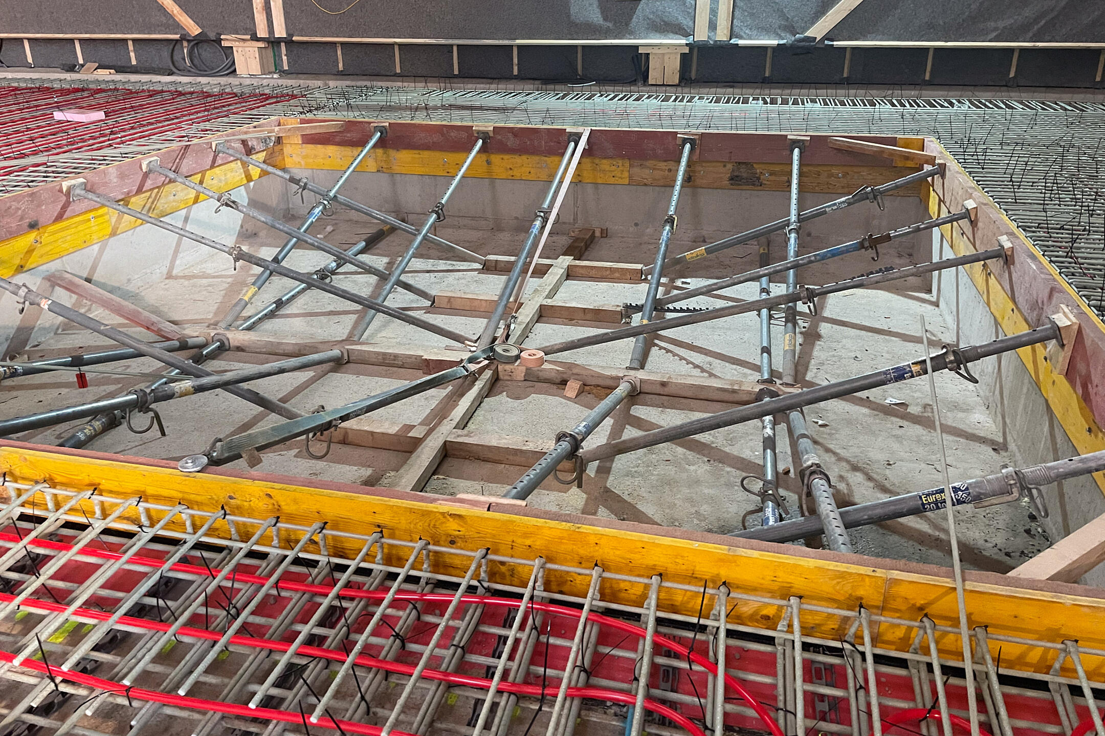 Detailansicht einer Baustelle im Innenbereich mit Schalung, Aussteifungen, GFK-Bewehrung und roten Schläuchen zur Vorbereitung einer Betonbodenplatte.
