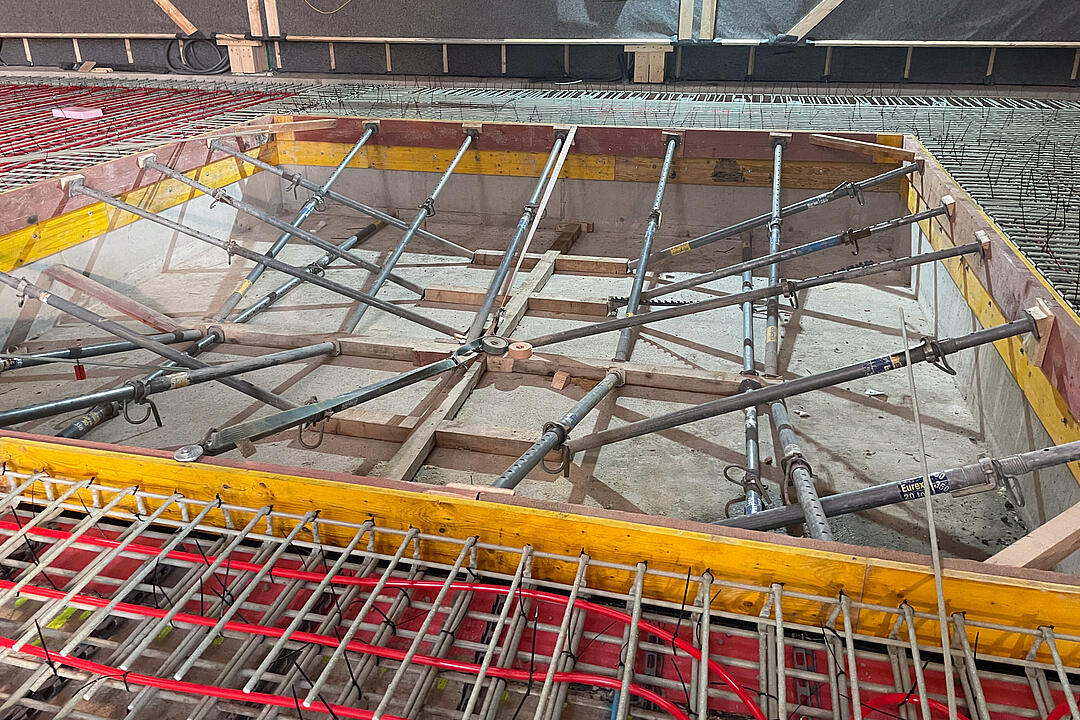 Detailansicht einer Baustelle im Innenbereich mit Schalung, Aussteifungen, GFK-Bewehrung und roten Schläuchen zur Vorbereitung einer Betonbodenplatte.