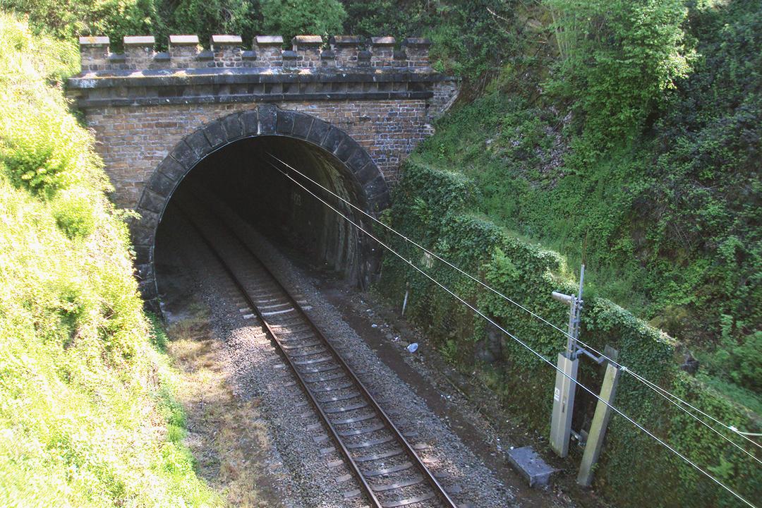 Bahntunnel aus Stein mit elektrifizierten Gleisen, umgeben von bewachsenem, grünem Hügelgelände im Sonnenlicht