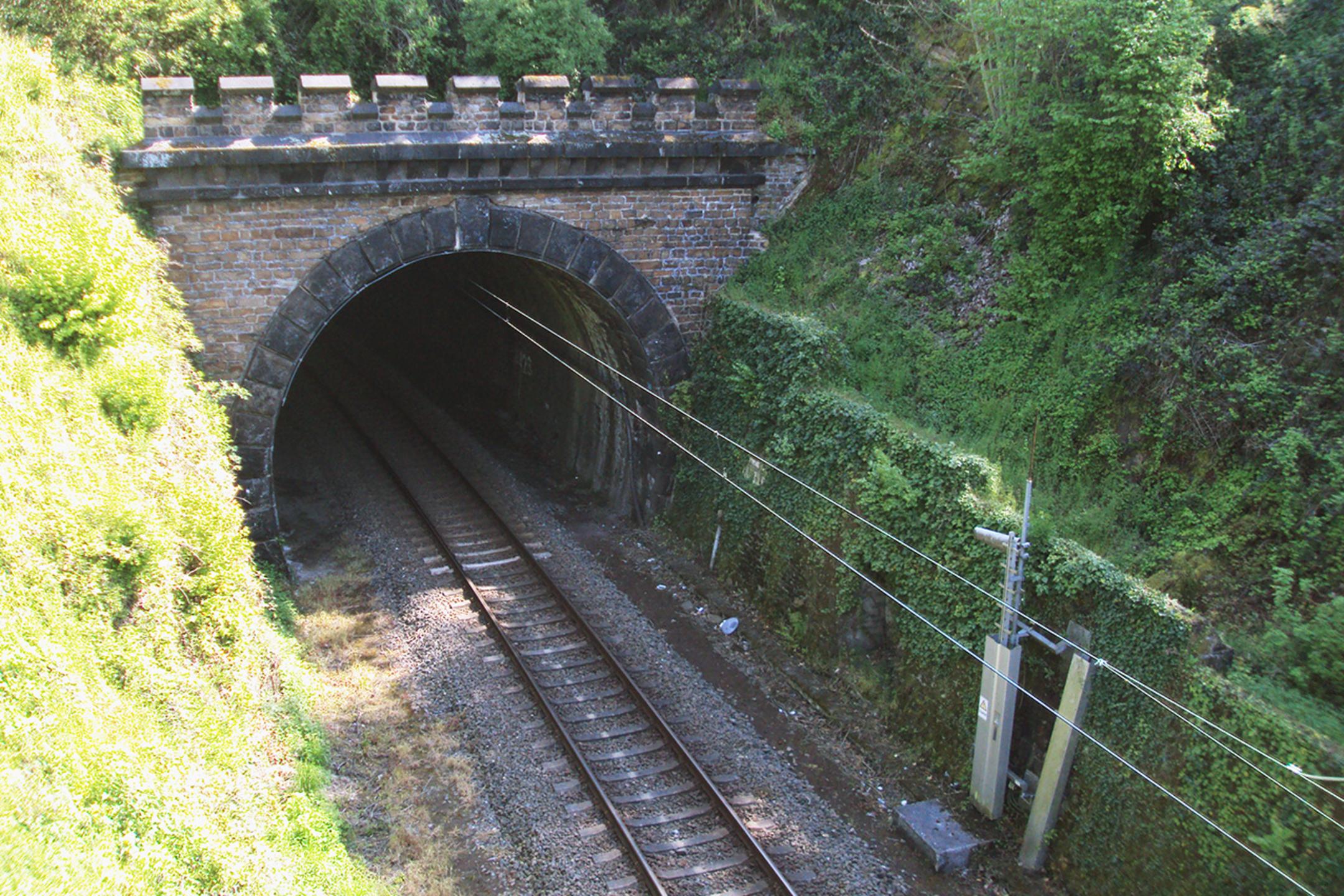 Bahntunnel aus Stein mit elektrifizierten Gleisen, umgeben von bewachsenem, grünem Hügelgelände im Sonnenlicht