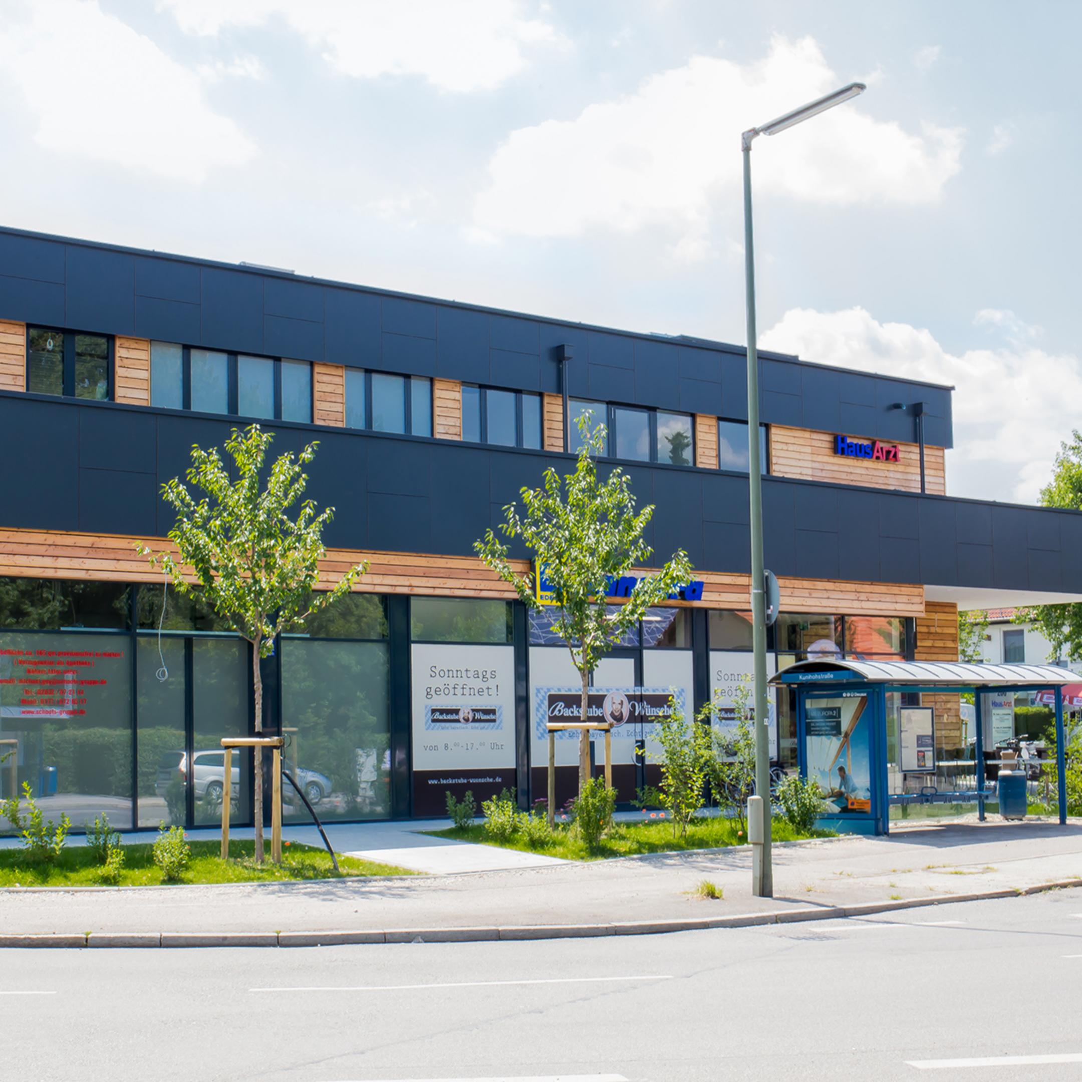 Modernes Gebäude mit großen Fenstern und Bushaltestelle davor, Firmenschilder sichtbar, leicht bewölkter Himmel