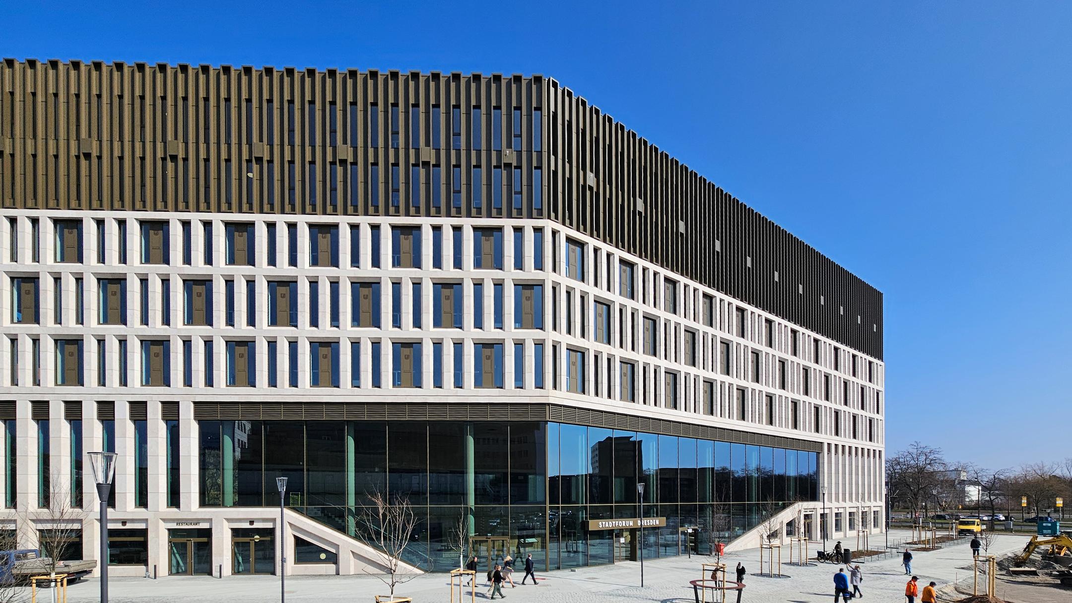 Gebäude vor blauem Himmel, im Vordergrund Menschen, Autos und Bäume