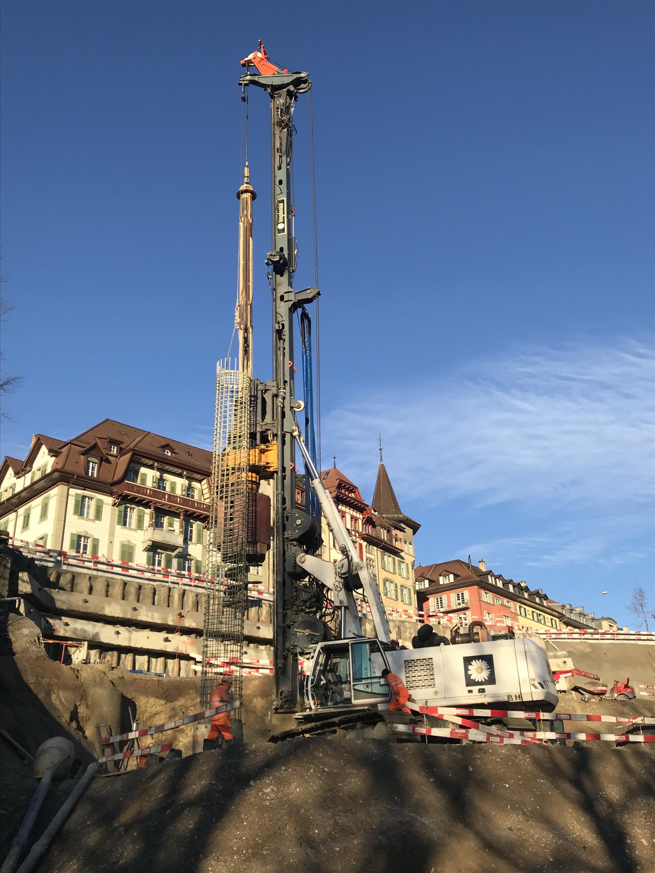 Baustelle mit Maschine und Bewehrungskorb, der in die Erde gelassen wird, vor mehrstöckigen Gebäuden bei klarem, blauem Himmel