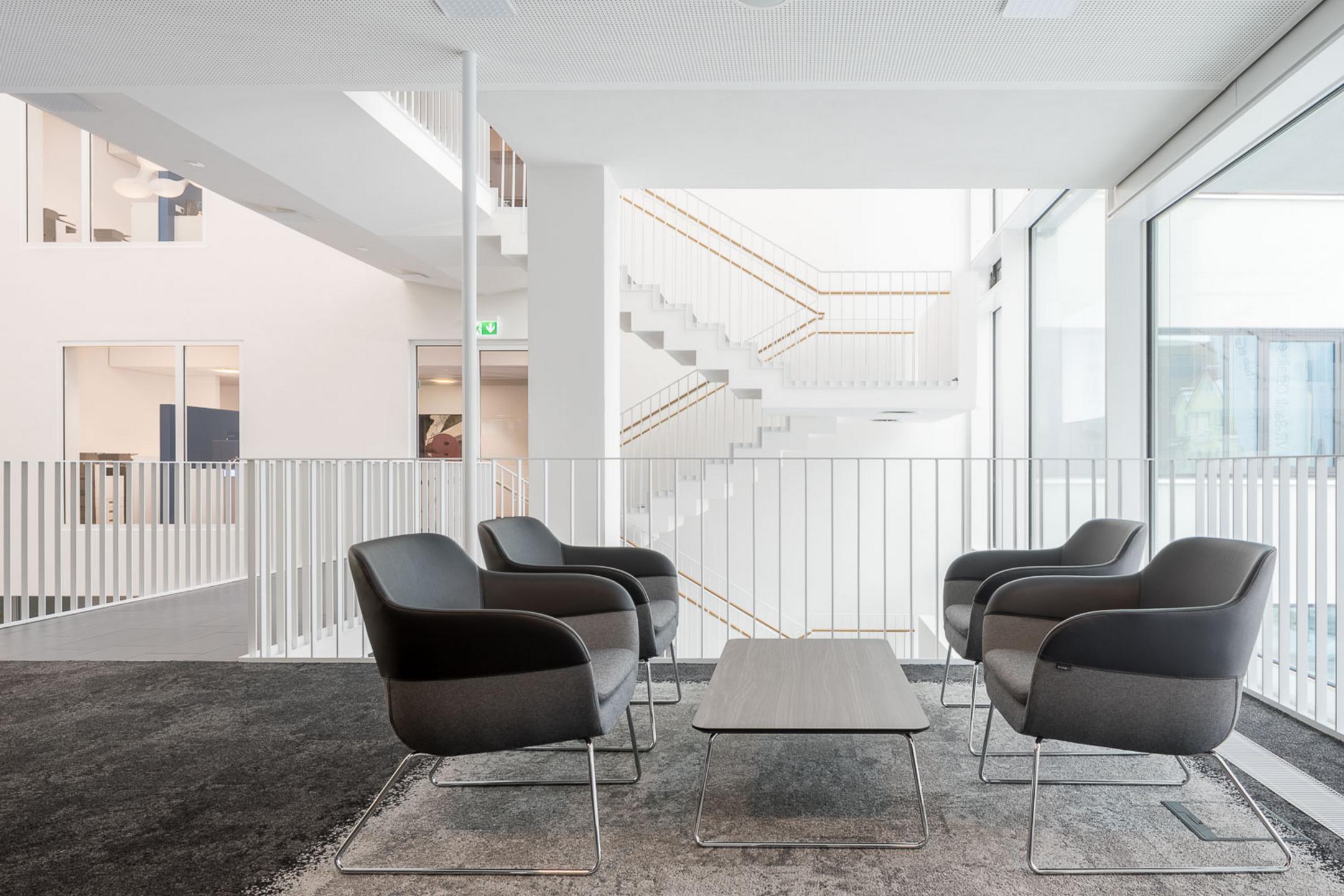 Moderne Lounge mit vier schwarzen Sesseln um einen Tisch, helle Treppe und große Fenster im Hintergrund