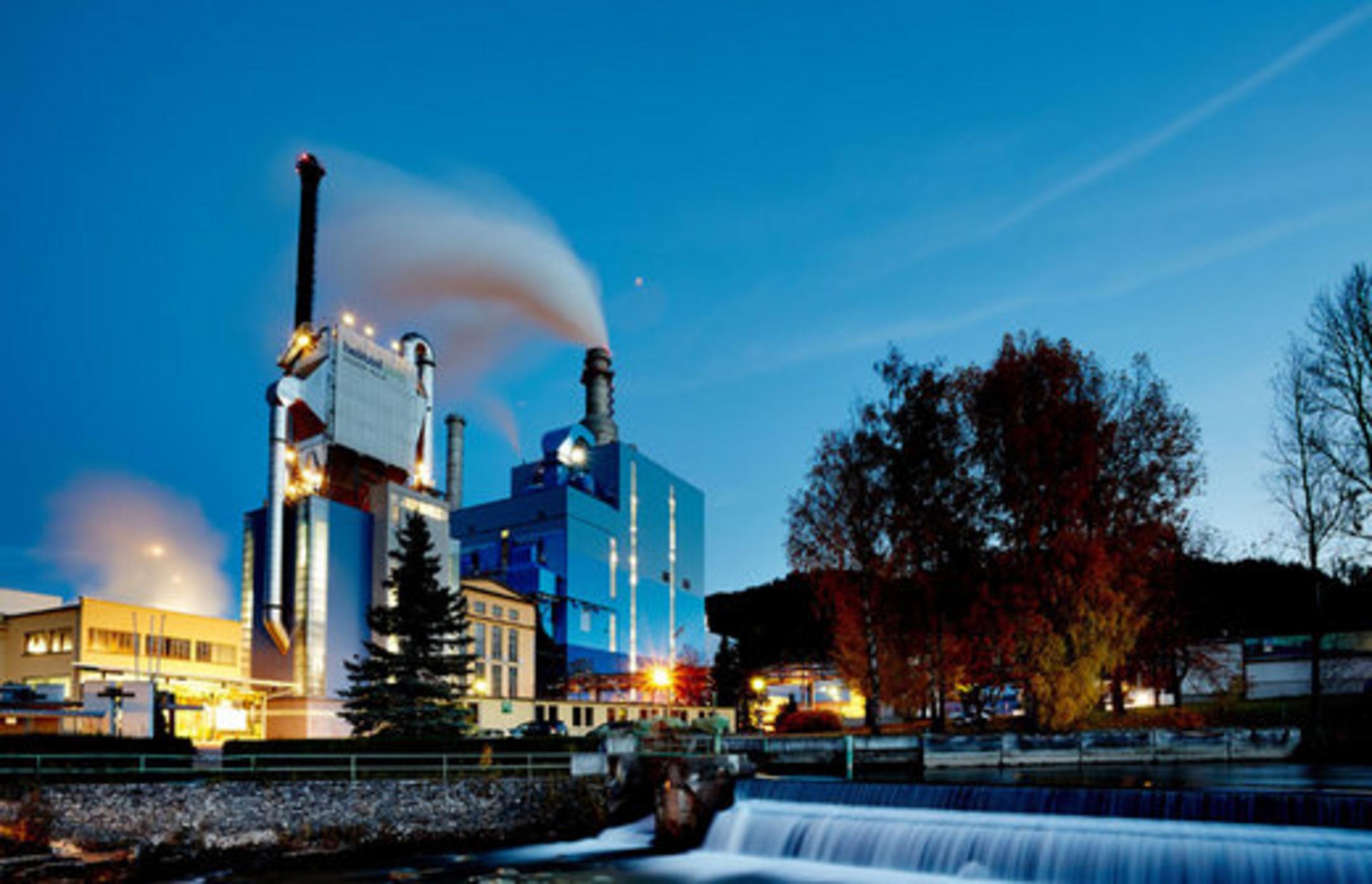Industrieanlage mit rauchenden Schornsteinen bei Dämmerung, nahe Wasserfall und umgeben von Natur und Gebäuden
