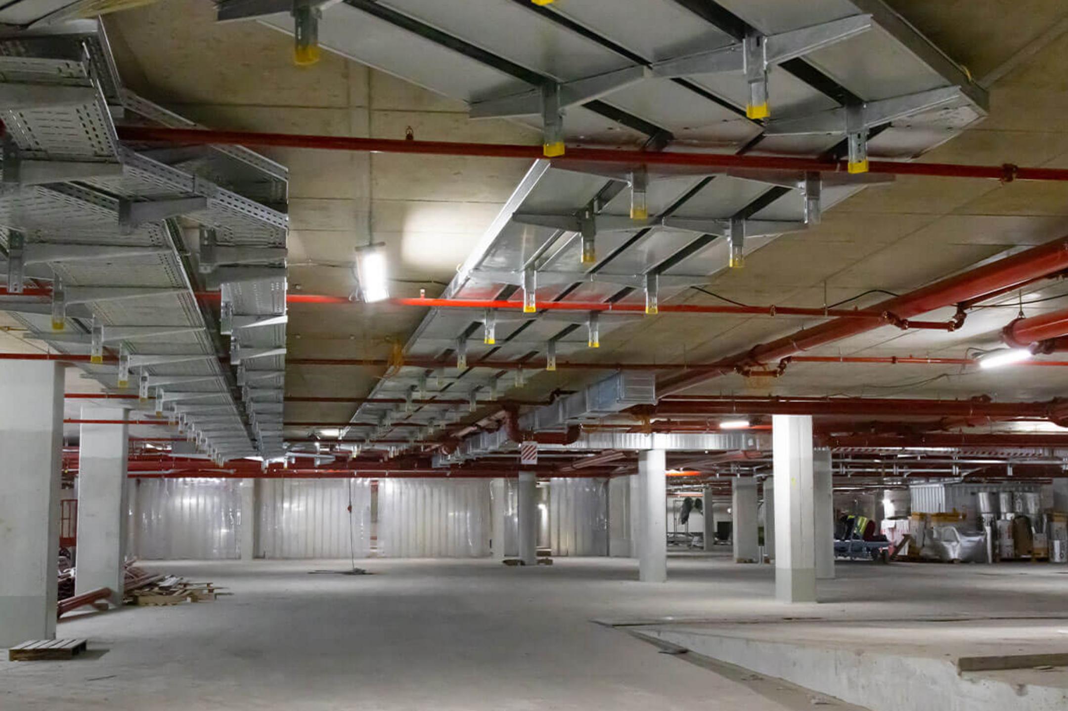 Tiefgarage im Rohbau mit an der Decke befestigten Konsolen und Kabelrinnen, dazwischen befinden sich rote Rohrleitungen