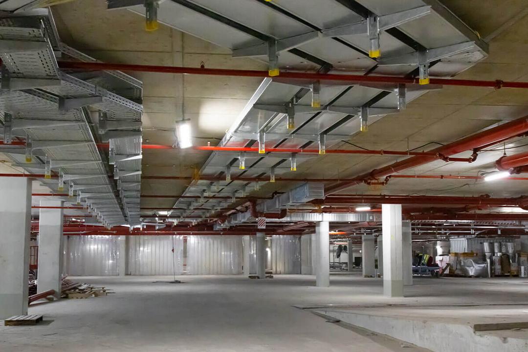Tiefgarage im Rohbau mit an der Decke befestigten Konsolen und Kabelrinnen, dazwischen befinden sich rote Rohrleitungen