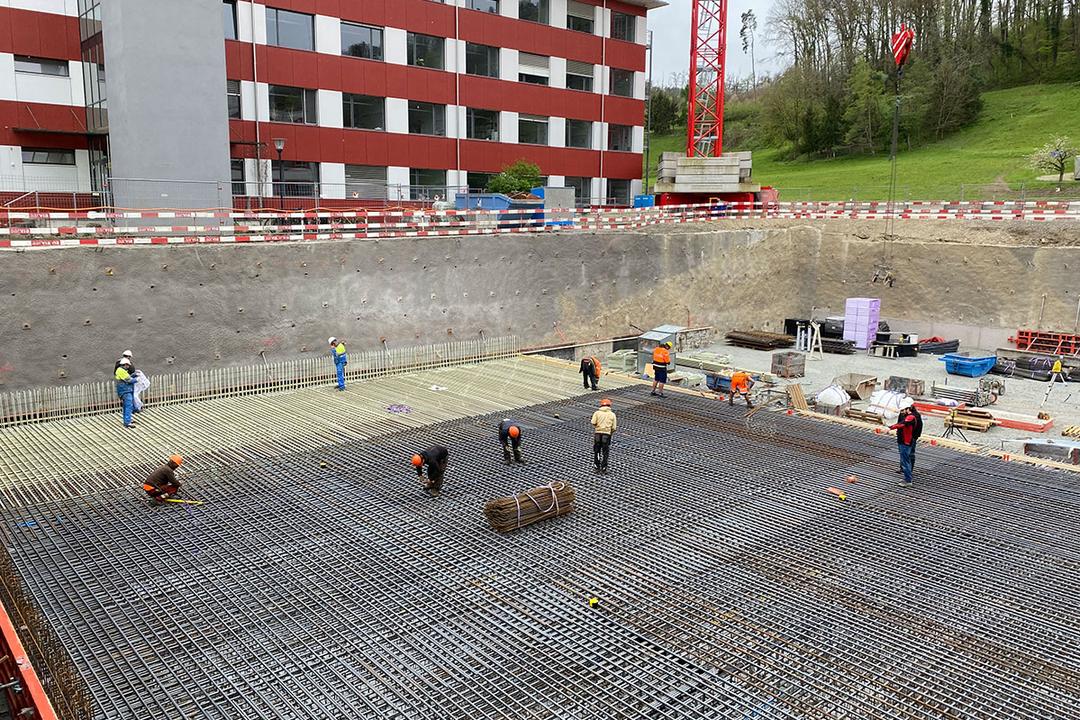 Bauarbeiter arbeiten an einem großen Baustellenfundament mit Glasfaserkunststoffarmierung in einer Baugrube vor einem roten Gebäude