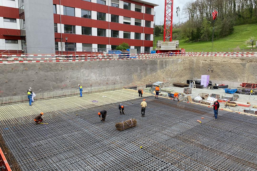 Bauarbeiter arbeiten an einem großen Baustellenfundament mit Glasfaserkunststoffarmierung in einer Baugrube vor einem roten Gebäude