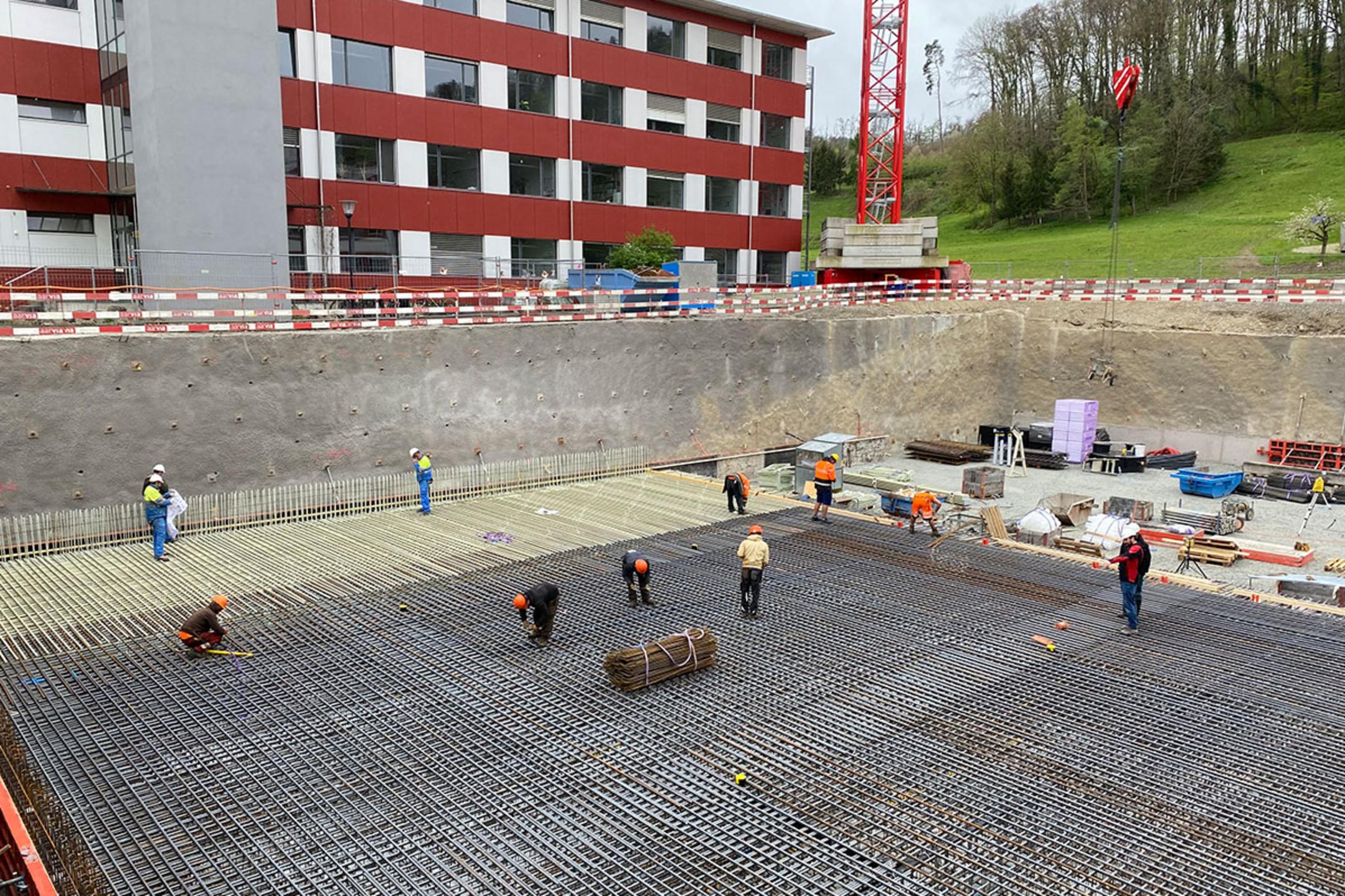Bauarbeiter arbeiten an einem großen Baustellenfundament mit Glasfaserkunststoffarmierung in einer Baugrube vor einem roten Gebäude