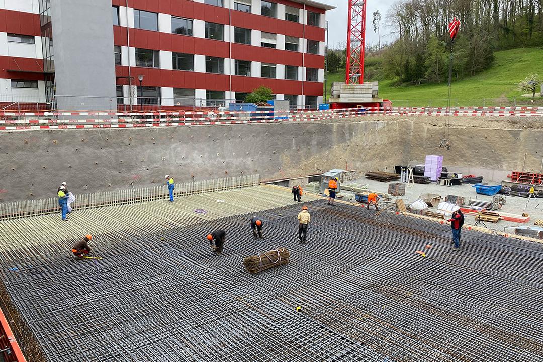 Bauarbeiter arbeiten an einem großen Baustellenfundament mit Glasfaserkunststoffarmierung in einer Baugrube vor einem roten Gebäude
