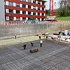 Bauarbeiter arbeiten an einem großen Baustellenfundament mit Glasfaserkunststoffarmierung in einer Baugrube vor einem roten Gebäude