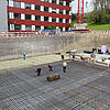 Bauarbeiter arbeiten an einem großen Baustellenfundament mit Glasfaserkunststoffarmierung in einer Baugrube vor einem roten Gebäude
