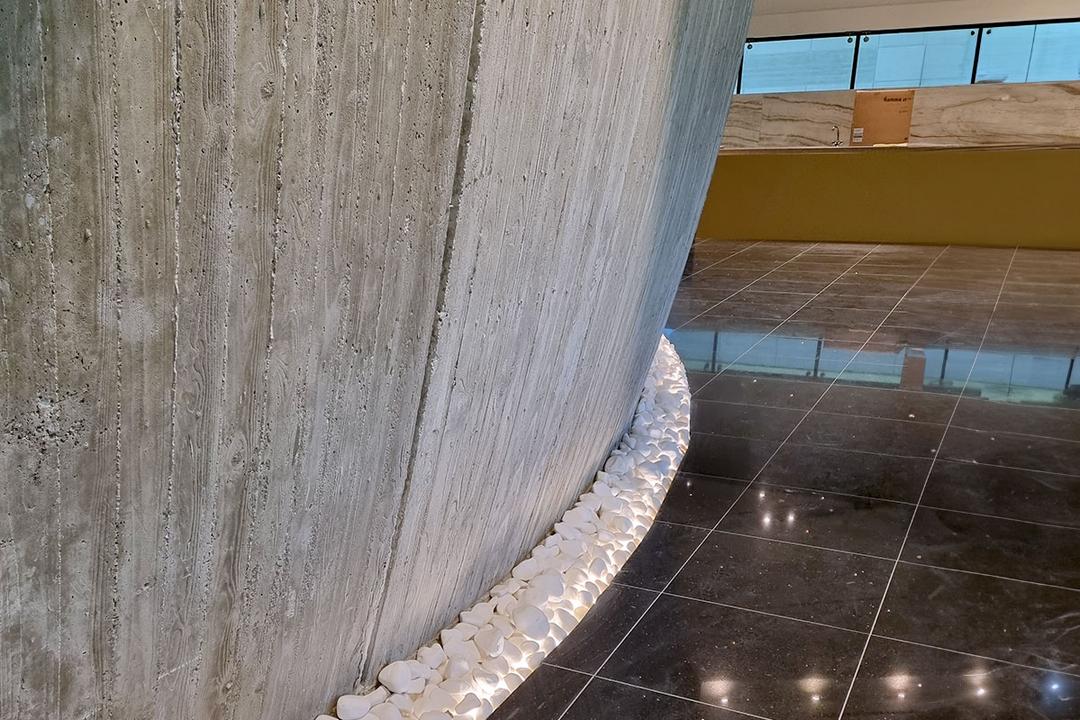 Gebogener Sichtbeton mit LED-beleuchteten weißen Ziersteinen an der Basis, daneben glänzender schwarzer Fliesenboden