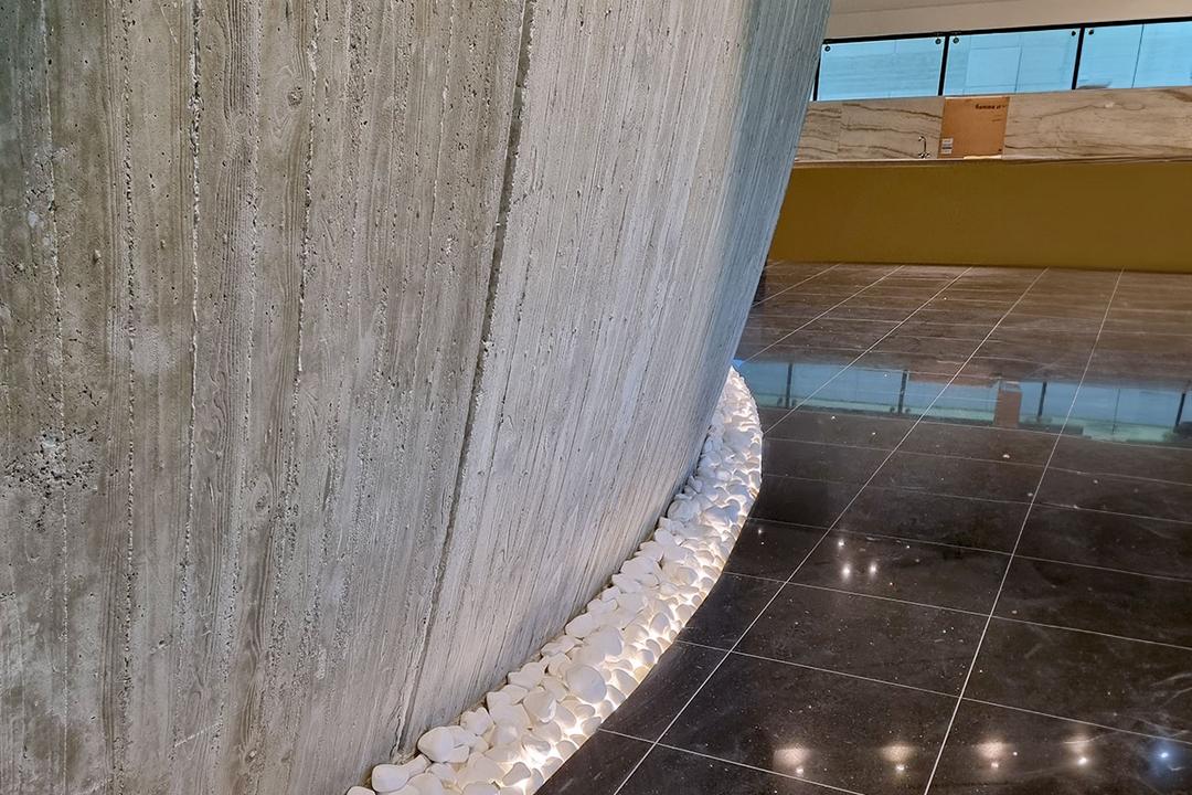 Gebogener Sichtbeton mit LED-beleuchteten weißen Ziersteinen an der Basis, daneben glänzender schwarzer Fliesenboden