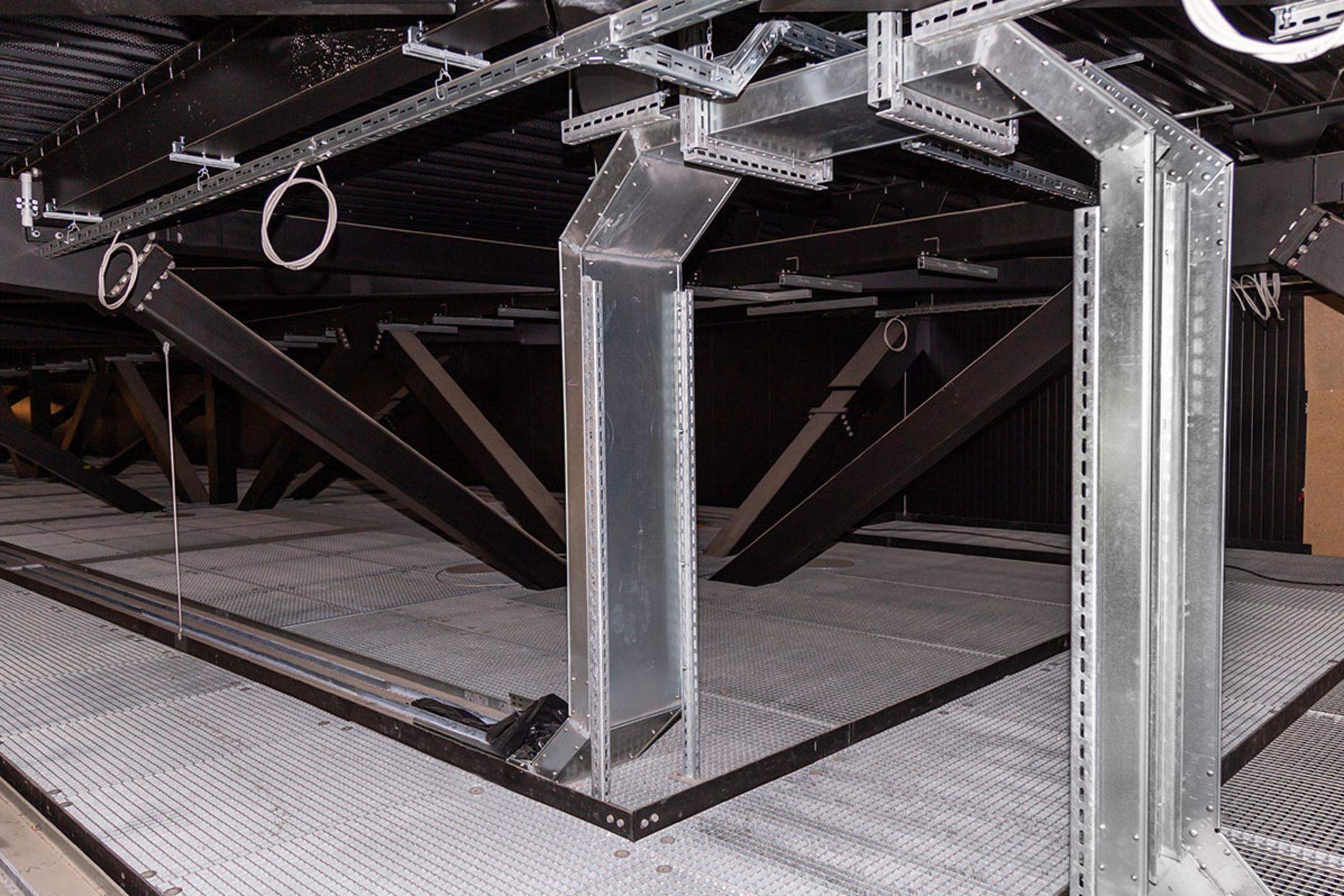 Vertikal verlaufende Kabelrinnen in einem Technikraum mit Gitterboden und dunkler Decke