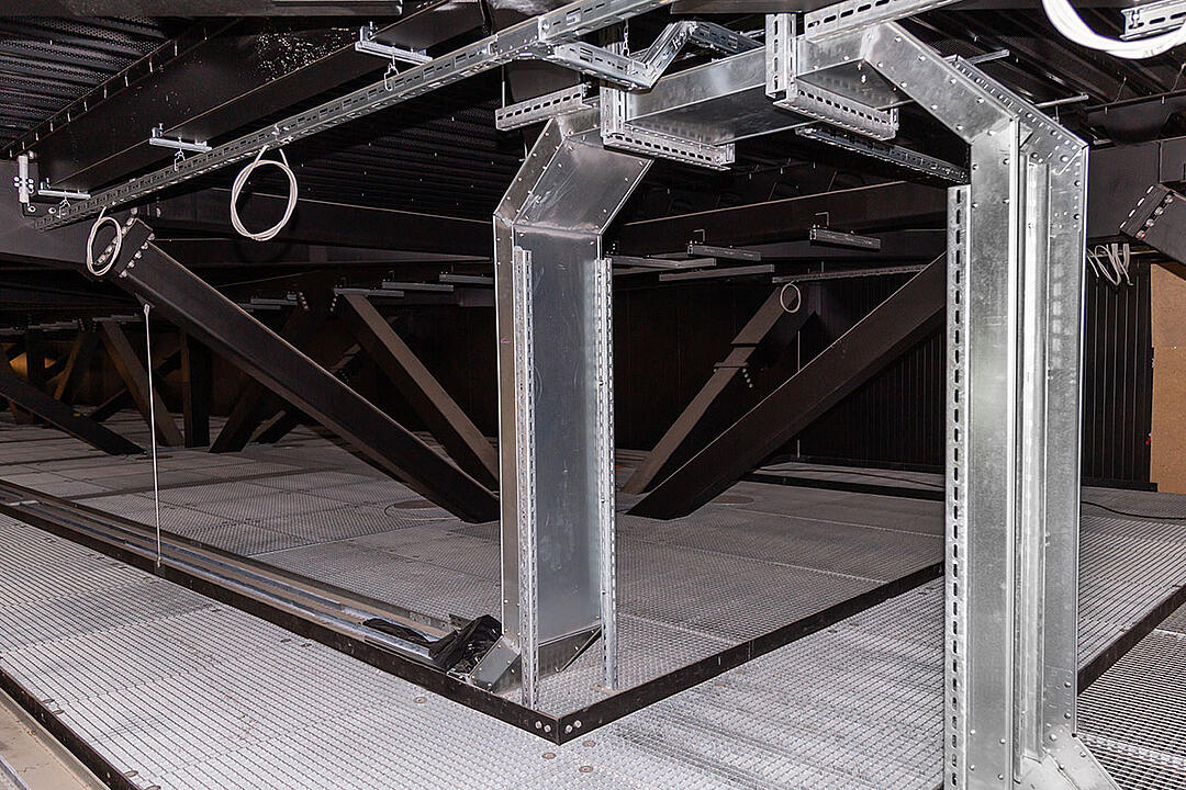 Vertikal verlaufende Kabelrinnen in einem Technikraum mit Gitterboden und dunkler Decke
