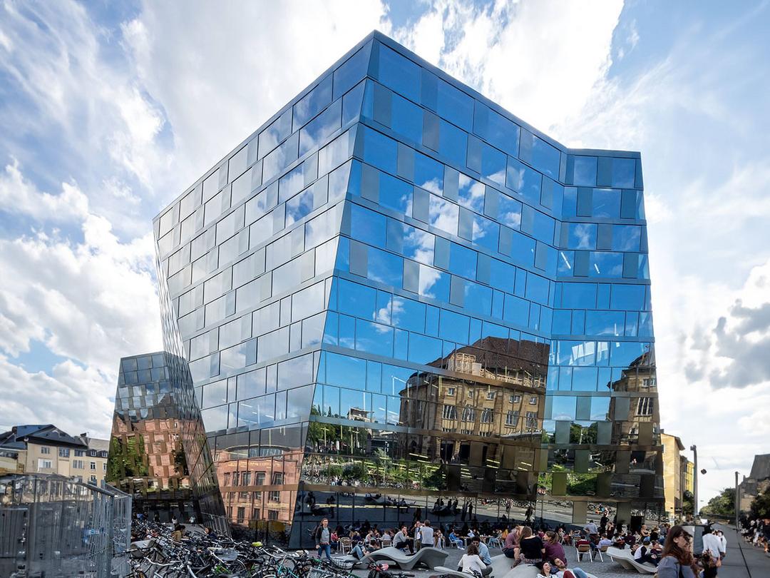 Modernes Gebäude mit spiegelnder Glasfassade, die Himmel und umliegende Architektur reflektiert, davor viele Menschen und Fahrräder
