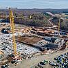 Blick von oben auf die Baustelle, viele Container und Baugerüste sind erkennbar, ringsherum sind Bäume und Felder