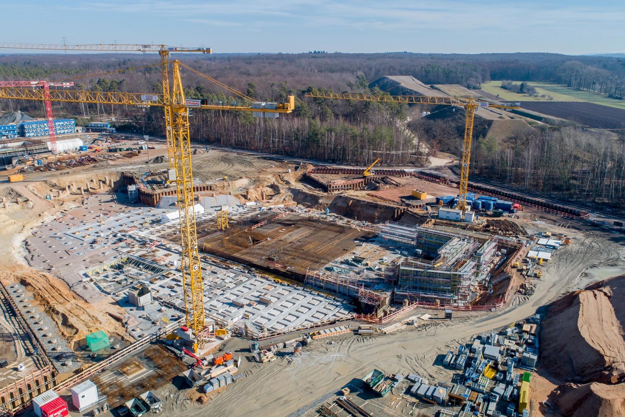 Blick von oben auf die Baustelle, viele Container und Baugerüste sind erkennbar, ringsherum sind Bäume und Felder