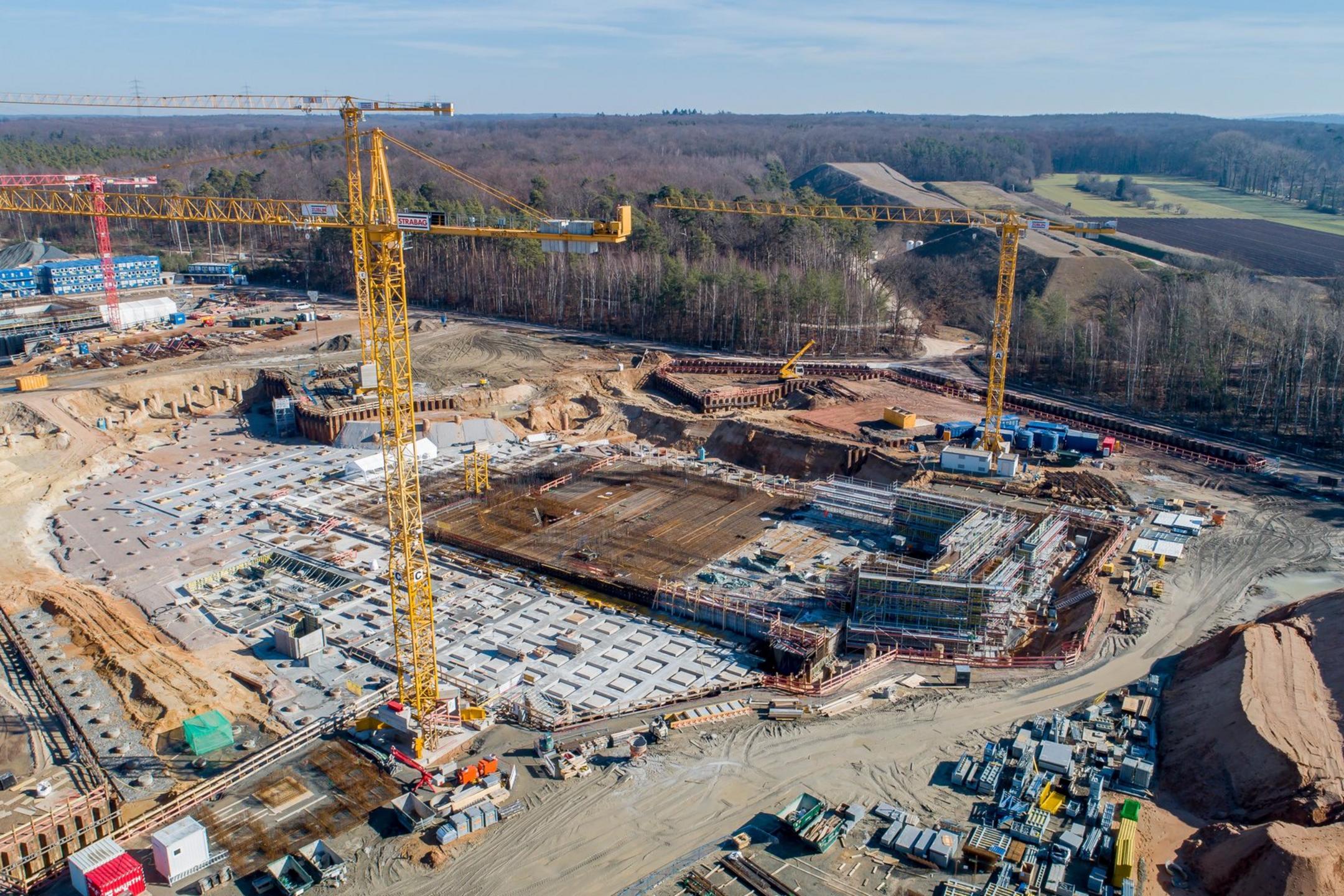 Blick von oben auf die Baustelle, viele Container und Baugerüste sind erkennbar, ringsherum sind Bäume und Felder